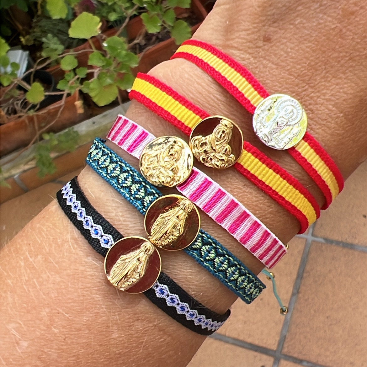 pulsera de cordón ajustable con medalla de la virgen del rocío, covadonga, milagrosa, del carmen y la milagrosa. es de acero hipoalergénico bañado en oro o plata. la cinta es de la bandera de españa, rosa, azul o negra. Es resistente al agua y apta para niños y hombre. 
